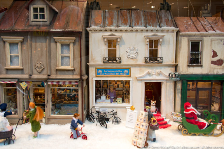 Miniature snowy street scene with shopfronts, tiny figures, bicycles, merchandise, and a red sleigh.