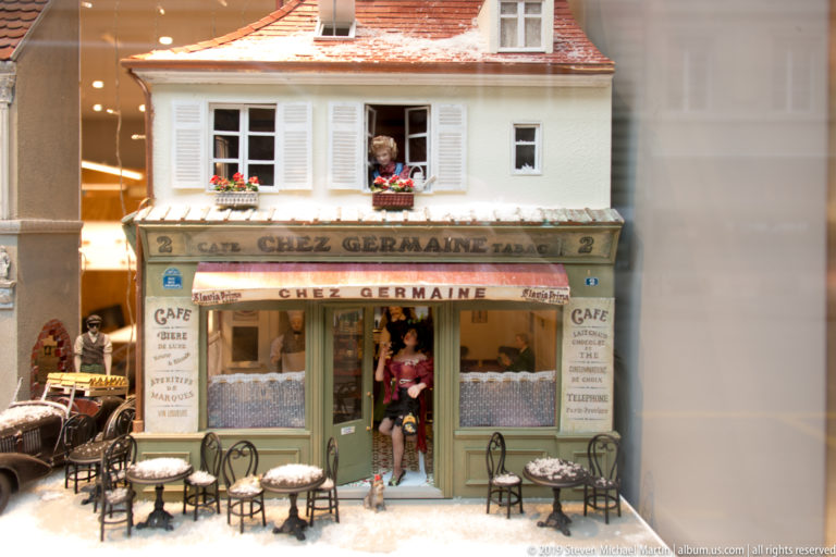 Miniature café façade labeled Chez Germaine with striped awnings, shutters, tiny tables, flowers, and small figures.