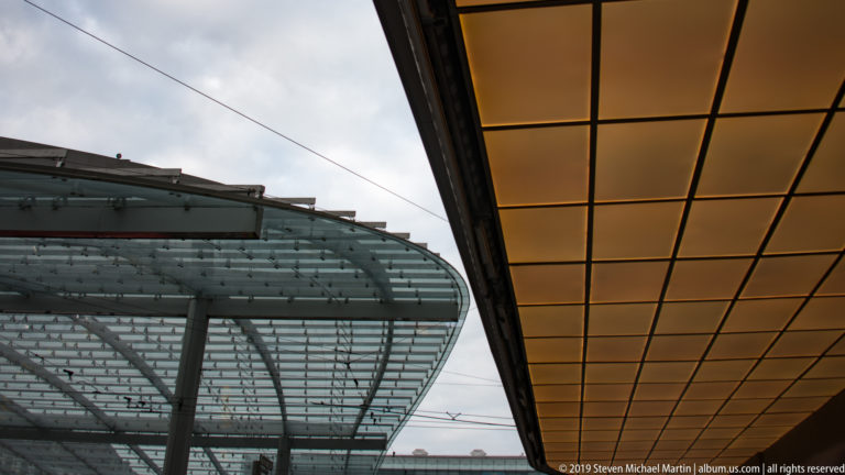 Upward view between a curved glass-and-steel canopy and an amber grid ceiling in Bern.