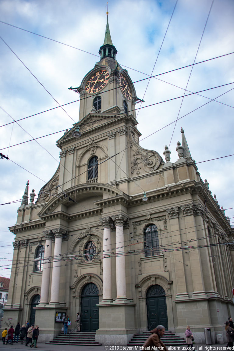 Baroque façade of Bern’s Church of the Holy Spirit with clock tower, green roof, columns, and overhead tram wires.