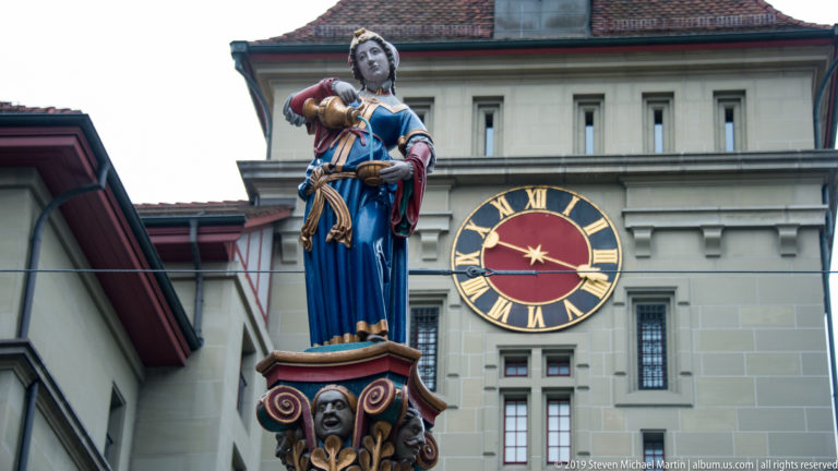 Polychromed female fountain figure in a blue gown standing before a large clock face in Bern.