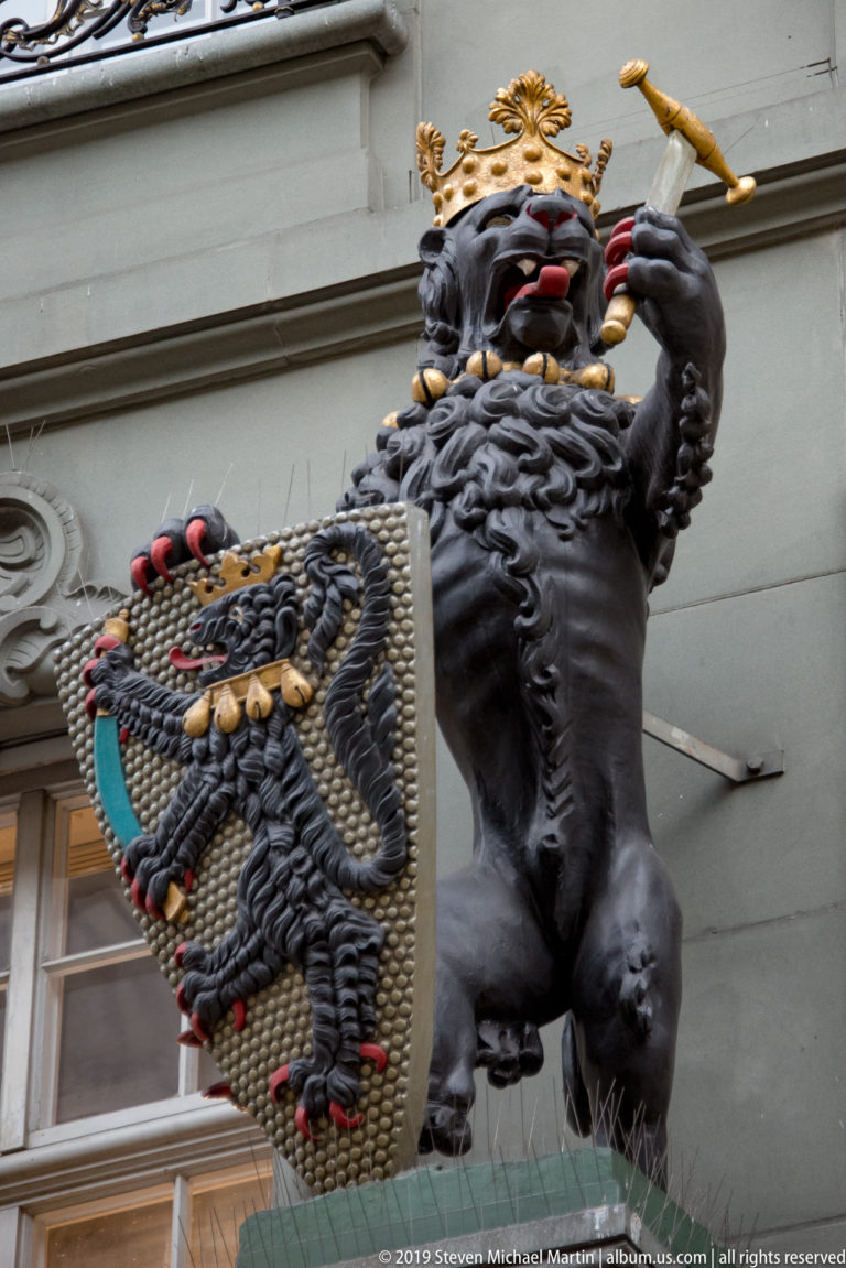 Large crowned black bear holding a staff and ornate heraldic shield on a Bern façade.
