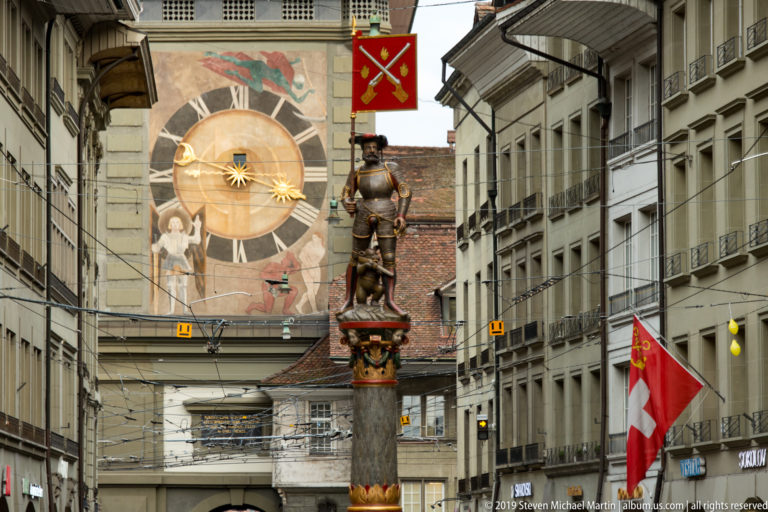 Armored fountain figure on a tall column aligned with a large painted clock on a Bern street.