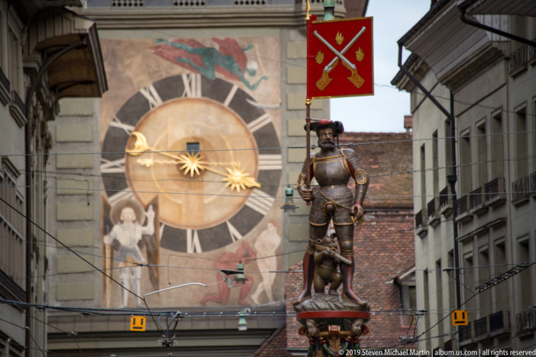 Armored fountain figure with red crossed-weapons banner and small bear, with a large clock in the background.