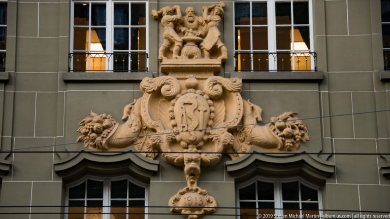Ornate stone cartouche with reclining figures, floral scrollwork, and a pendant dated 1915 between two windows.