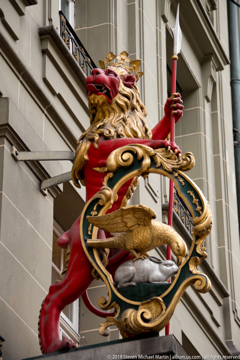 Red crowned lion holding a spear above a gilded cartouche containing a bird emblem.