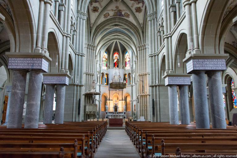 Central nave of Bern Minster with pointed arches, ribbed vaults, wooden pews, and stained glass toward the choir.
