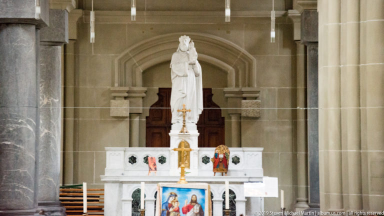 Decorated white side altar with crowned female statue, candles, cross, and painted devotional panel.