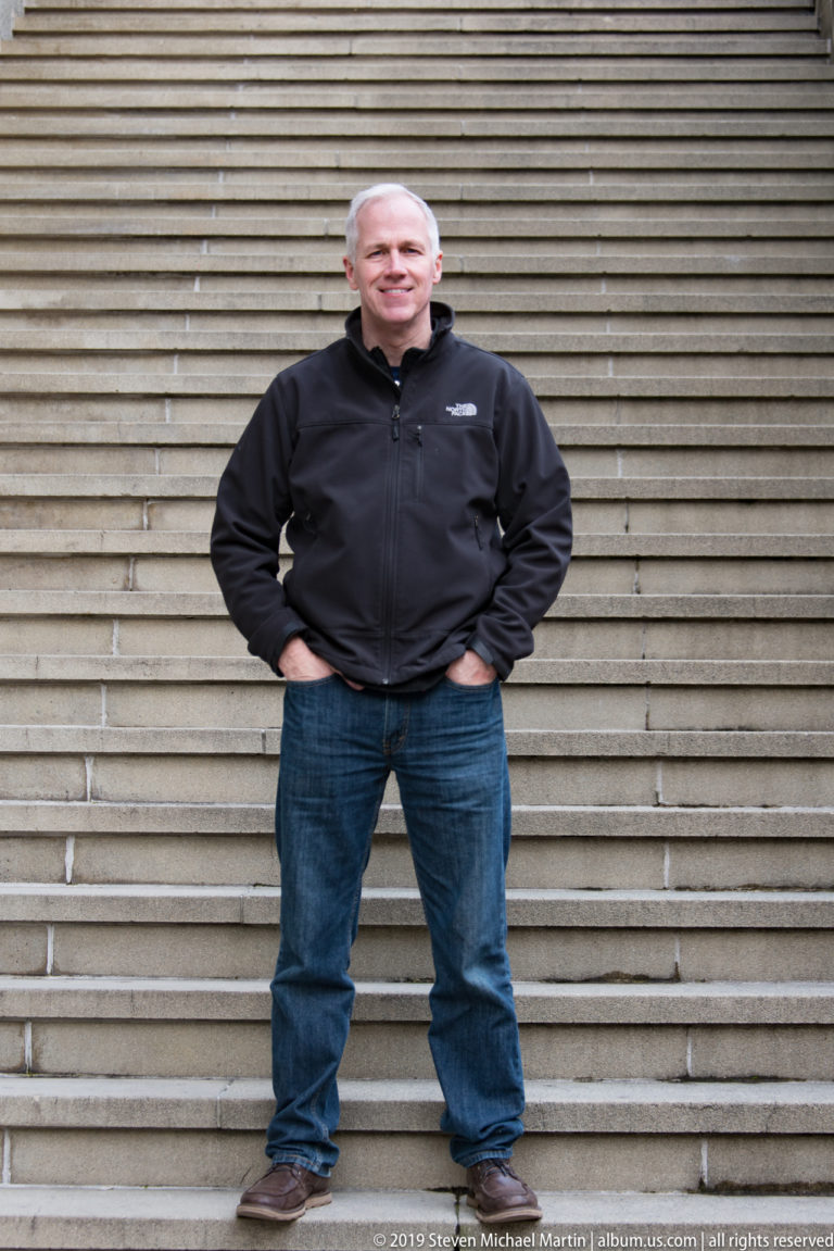 Man in black jacket and jeans standing centered on a broad flight of stone steps.