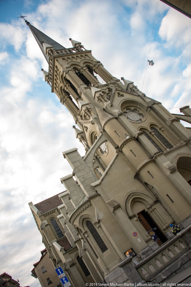 Steep low-angle view up Bern Minster’s Gothic façade and tower beneath a cloudy sky.