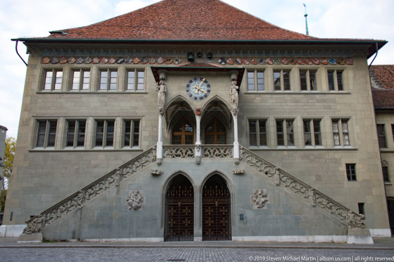 Front view of Bern Town Hall with twin exterior stairways, Gothic loggia, clock, heraldic shields, and red tiled roof.