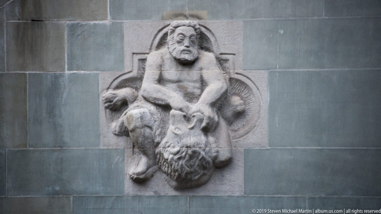 Weathered stone relief of a human figure positioned above a large grotesque animal-like head.