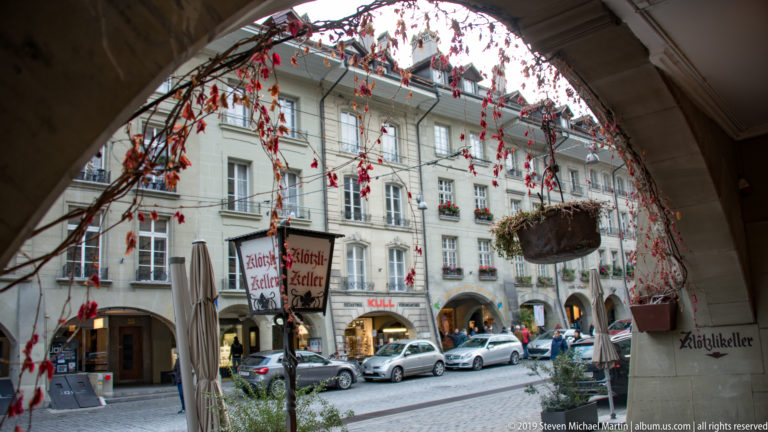 View through a Bern Old City arcade with red autumn vines toward shops, parked cars, and pale historic façades.
