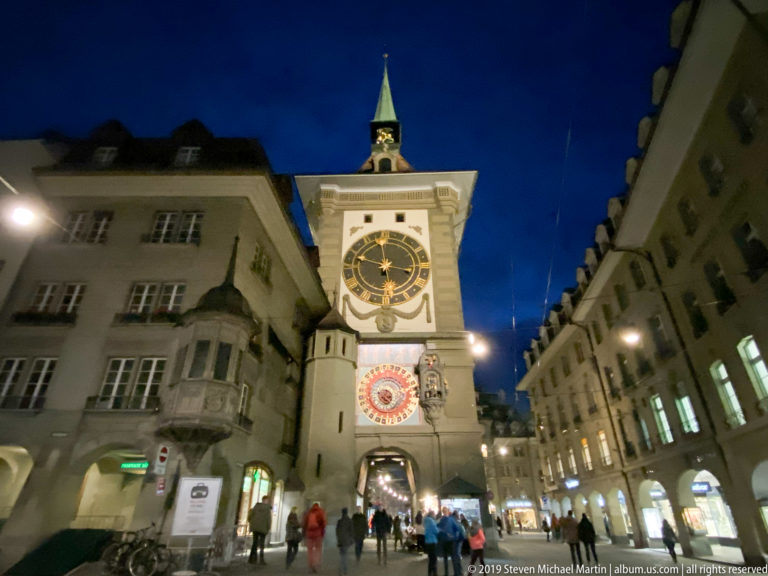 Bern’s illuminated Zytglogge at night with upper clock, astronomical dial, archway, and pedestrians below.