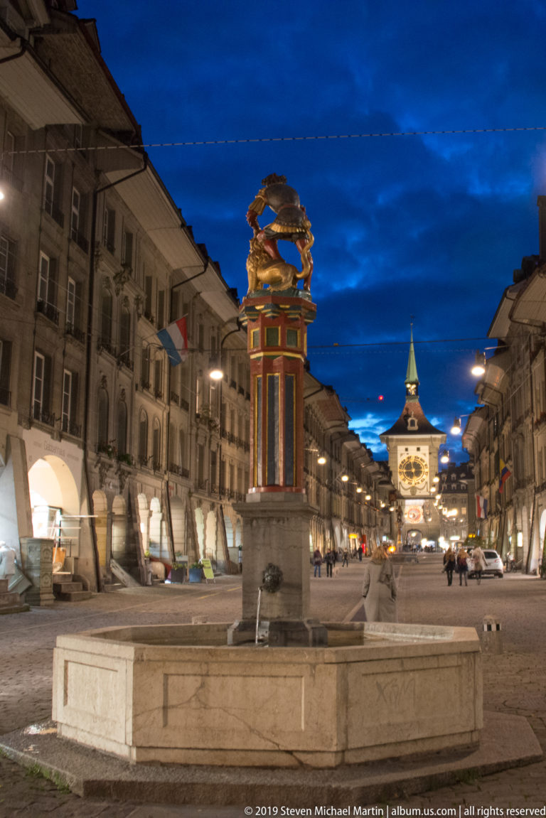 Polychromed historic fountain on a Bern street at night with the illuminated Zytglogge in the distance.