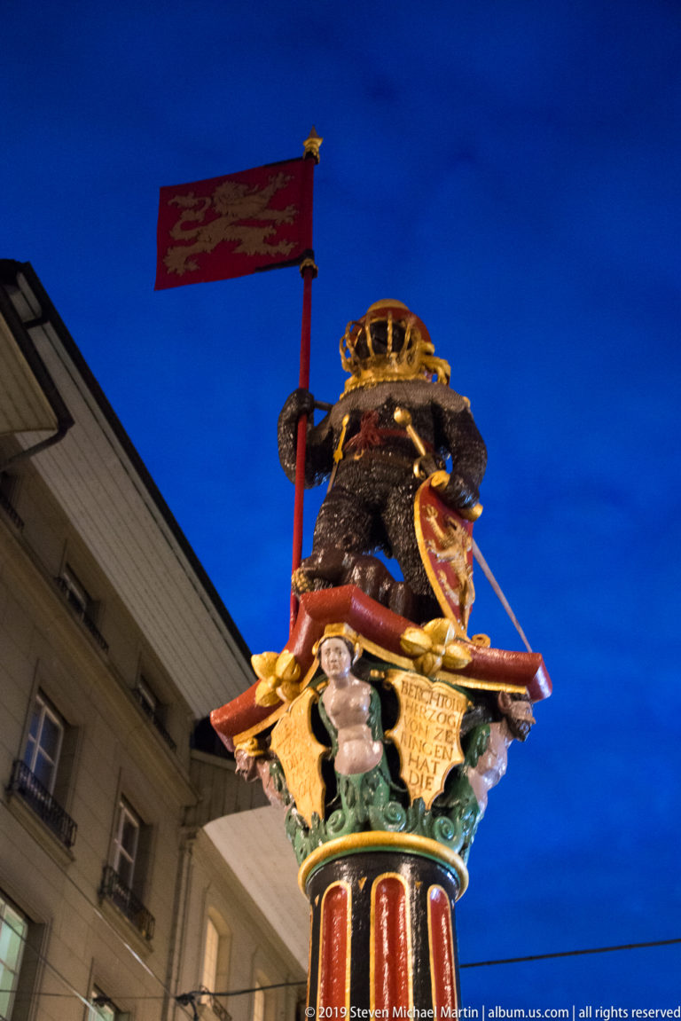 Low-angle night view of the armored bear and cub atop Bern’s Zähringerbrunnen, with a tall red banner.