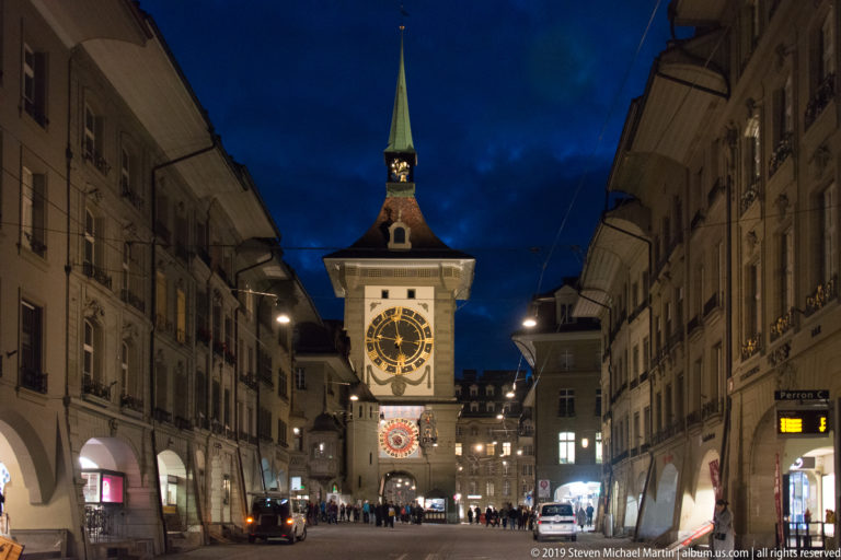 Frontal night view of Bern’s Zytglogge centered between arcaded buildings and lit shopfronts.