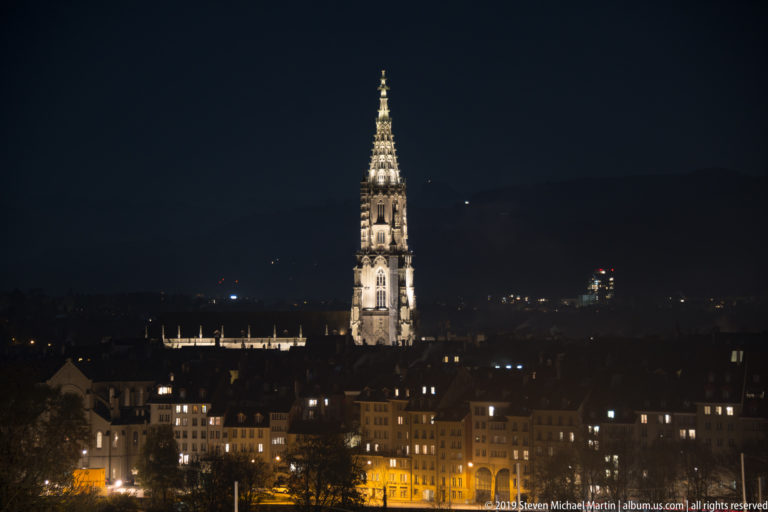 Illuminated Gothic tower of Bern Minster rising above dark Old City rooftops at night.