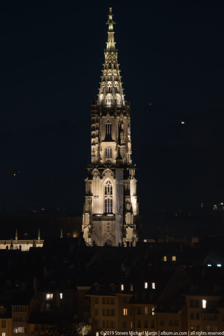 Illuminated Gothic tower and openwork spire of Bern Minster rising above dark rooftops at night.