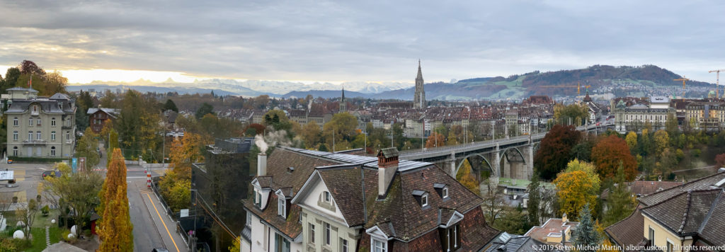 Elevated autumn view over Bern rooftops toward a long bridge, church spire, wooded hills, and distant snowy Alps.