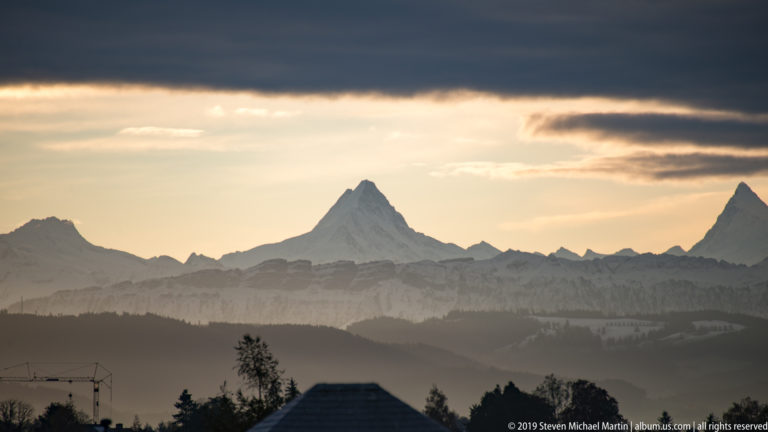 Layered silhouettes of the Bernese Alps at dusk beneath dark clouds and a glowing horizon.
