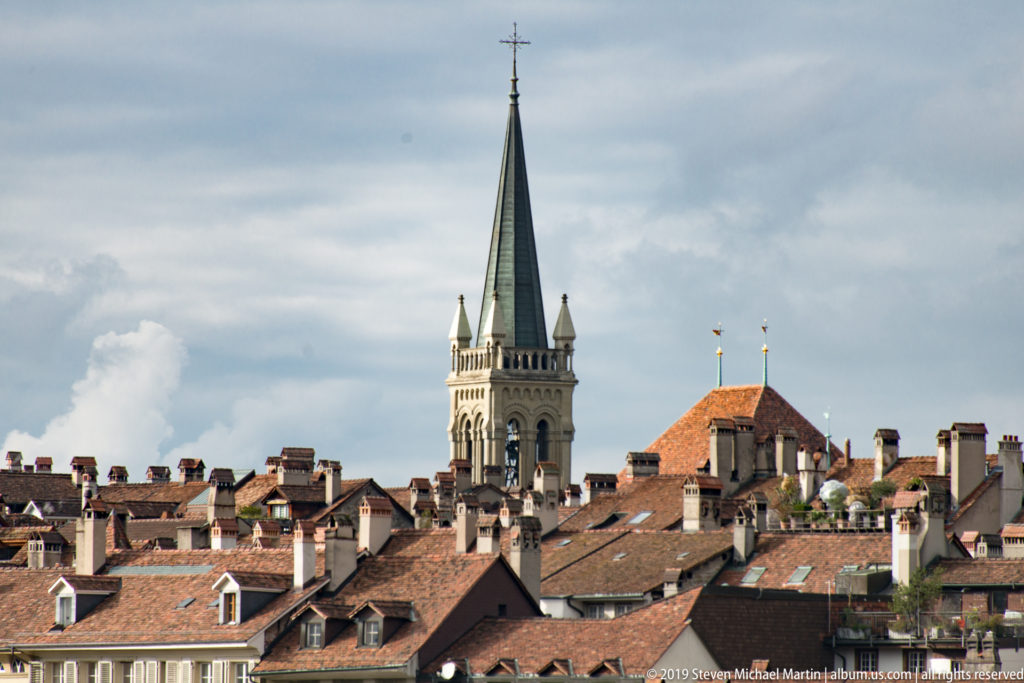 Slender green-spired church tower rising above tightly packed red-tiled roofs in Bern.