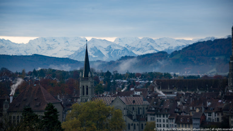 Church spire above Bern’s red-tiled rooftops with wooded hills and snowy Bernese Alps behind.