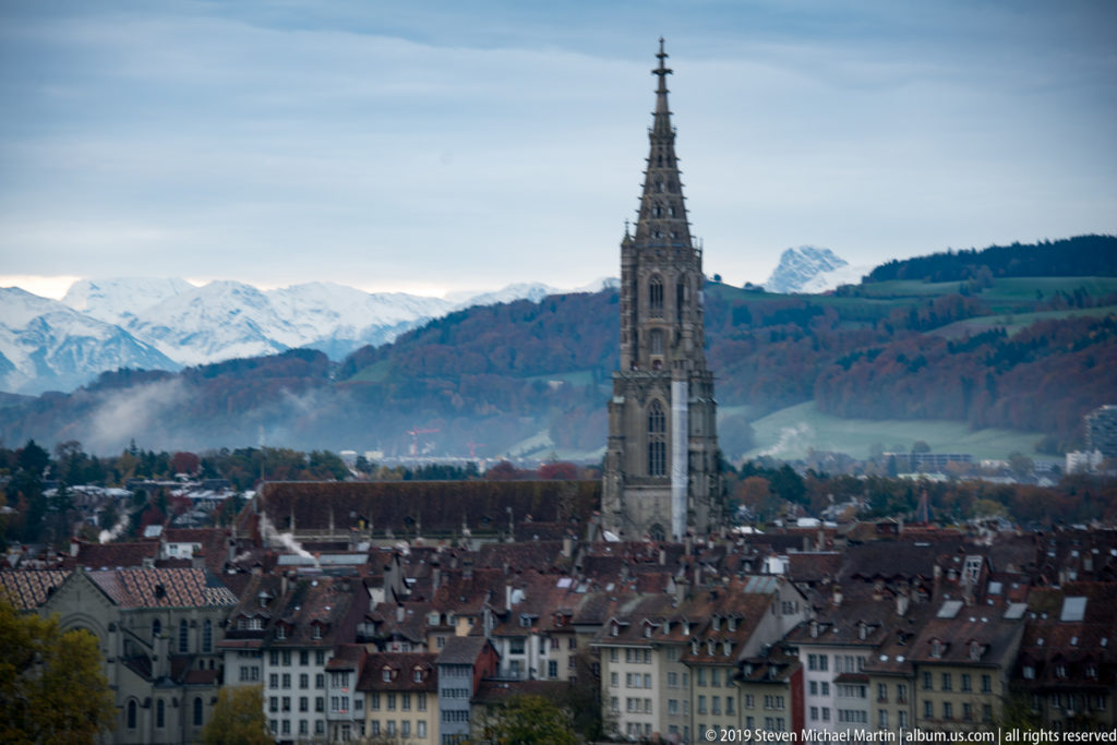 Bern Minster tower above dense Old City rooftops with wooded hills and snowy Bernese Alps in the background.