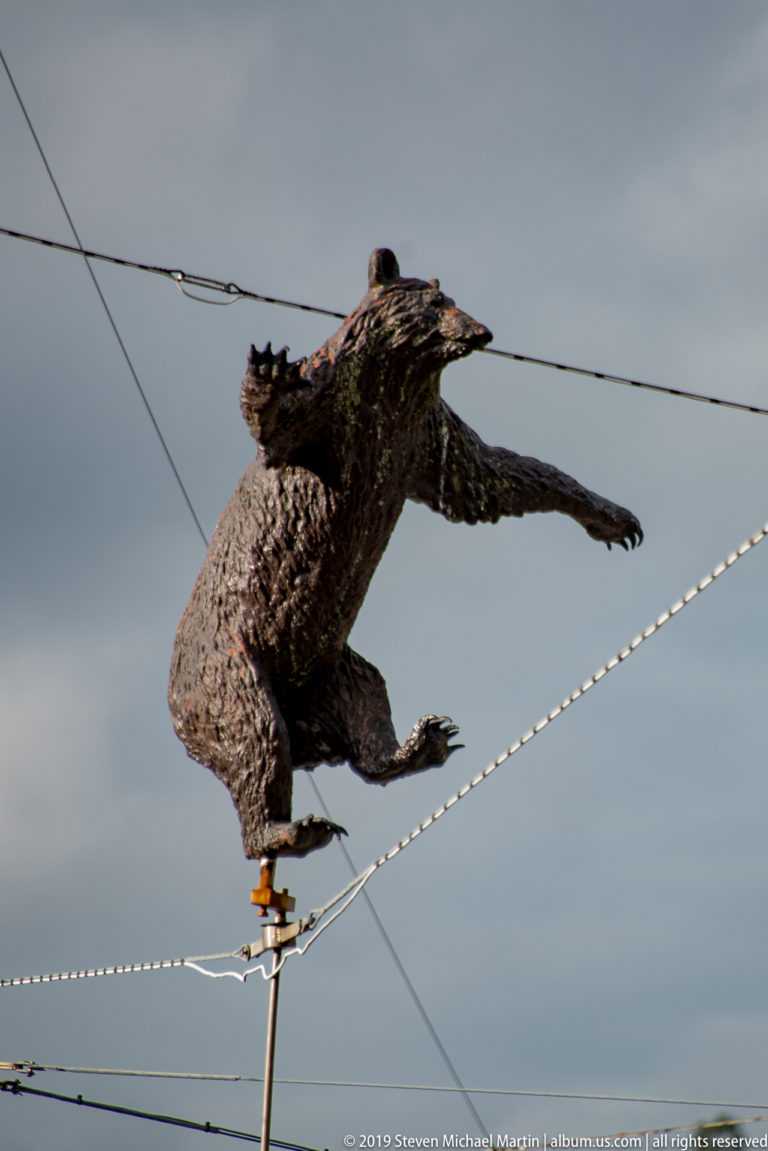 Dark bear sculpture with outstretched forelegs mounted above overhead transit wires.