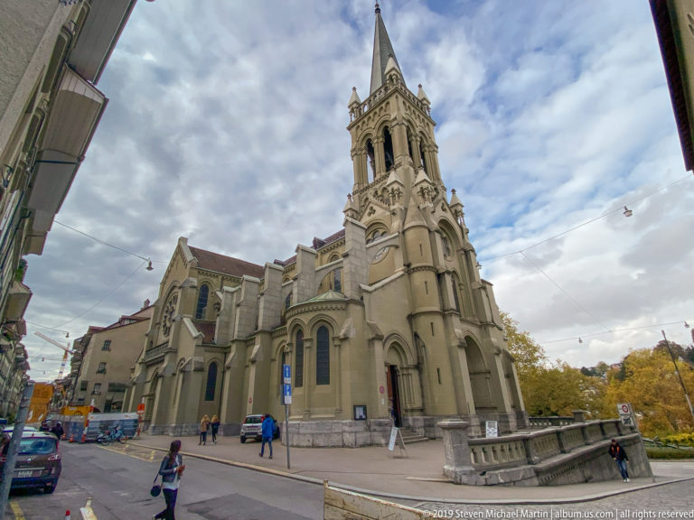Stone church with pointed arches and a tall spired tower at a Bern street corner.