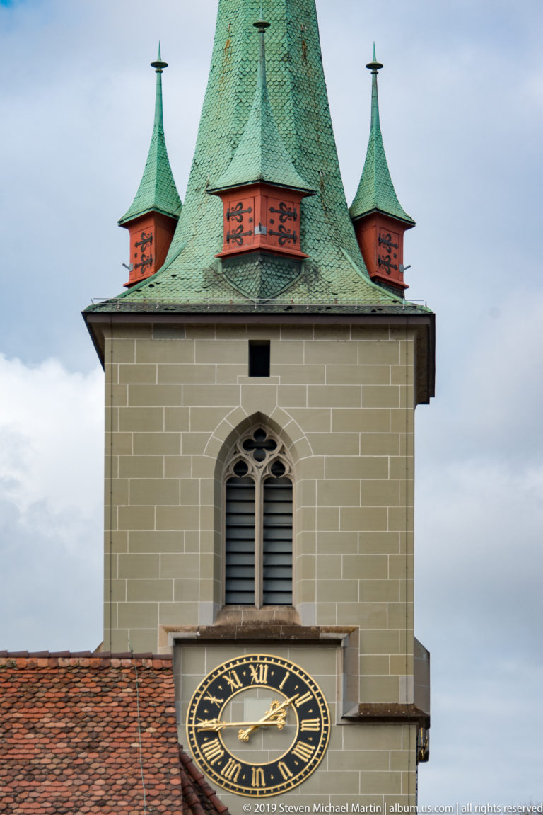 Black-and-gold clock on a square tower with a green roof and small corner turrets.