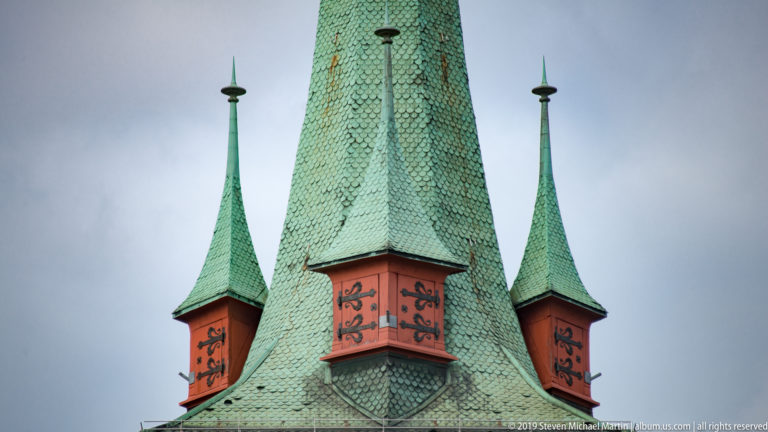 Green-patinated steep roof with two narrow spirelets and red-orange decorative dormer panels against a gray sky.