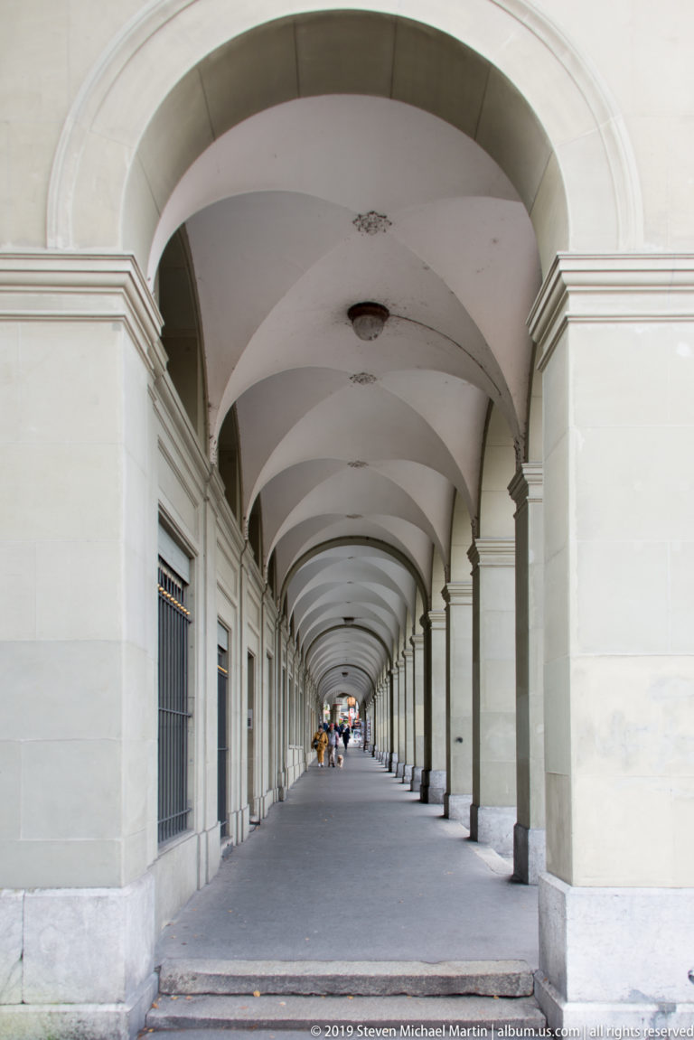 Long stone arcade with repeating arches and shallow vaults receding toward pedestrians in the distance.