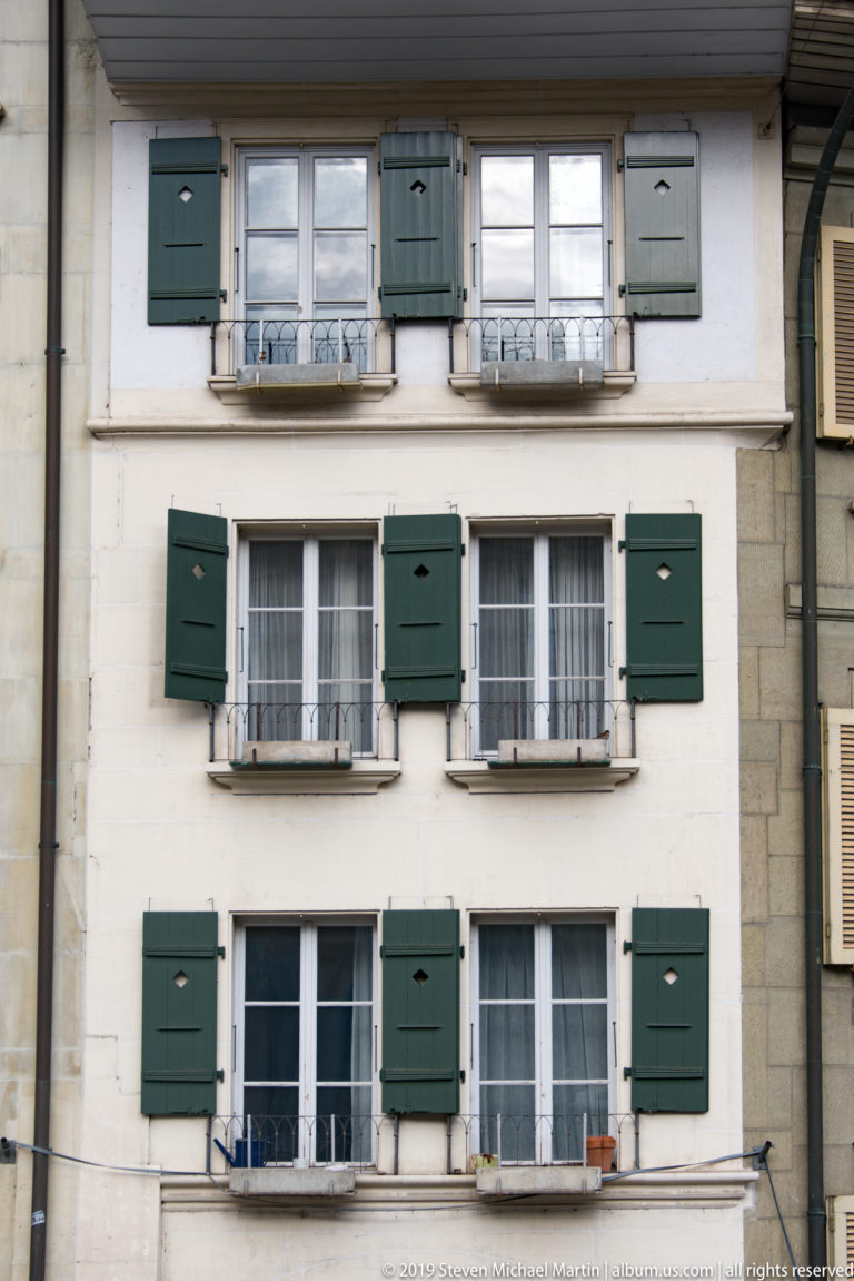 Pale historic facade with three rows of white windows, dark green shutters, and small wrought-iron guards.