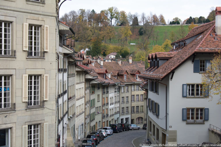 Narrow sloping Bern street lined with tiled-roof houses, parked cars, and a green hillside beyond.