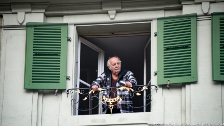 Older man leaning from an open window with green shutters and a decorative iron guard.
