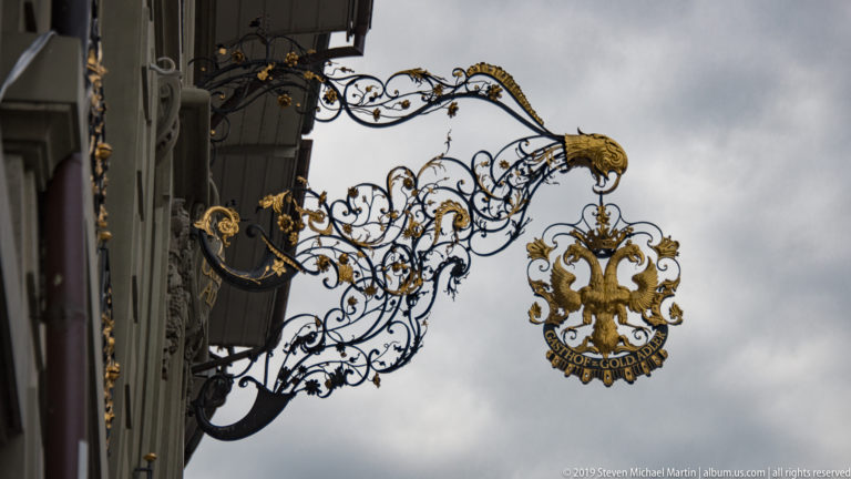 Ornate black wrought-iron hanging sign with gilded eagle motifs projecting from a building facade.