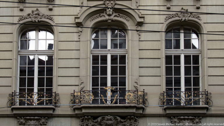 Three tall arched windows with ornate stone carvings and black-and-gold balcony railings on a historic facade.