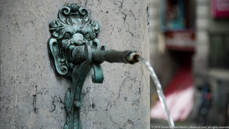 Close view of a weathered bronze grotesque mask and pipe pouring water beside pale stone.