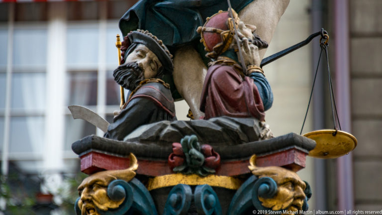 Painted subordinate figures clustered beneath the Justice statue, with part of a hanging balance scale at right.