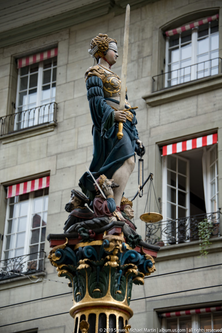 Full side view of blindfolded Justice with sword and scales atop a colorful fountain column in Bern.