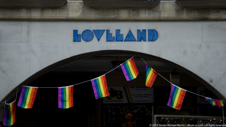Rainbow flags strung across a shadowed arched entrance beneath blue lettering on a stone facade.