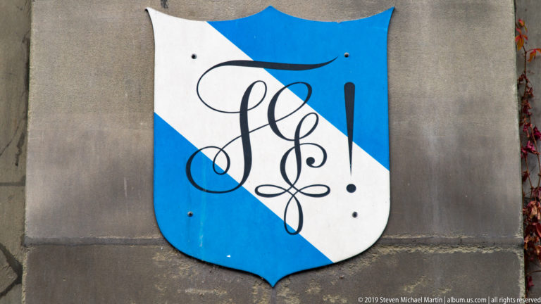 Blue-and-white shield plaque on stone wall with a large black calligraphic monogram.
