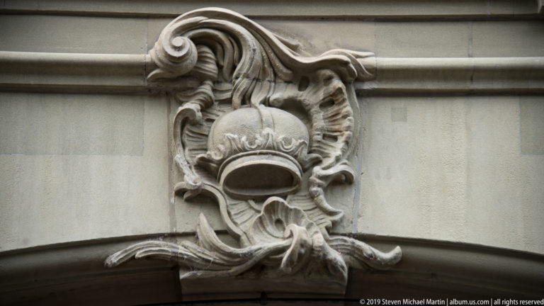 Carved stone helmet surrounded by curling scrollwork above a curved architectural opening.