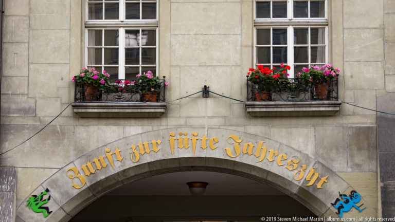 Stone arched entrance with gilded German lettering, flower-filled windows, and small painted heraldic animals.