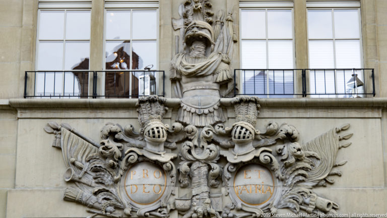 Large carved heraldic composition with helmets, scrolls, paired motto shields, and a projecting figure between windows.