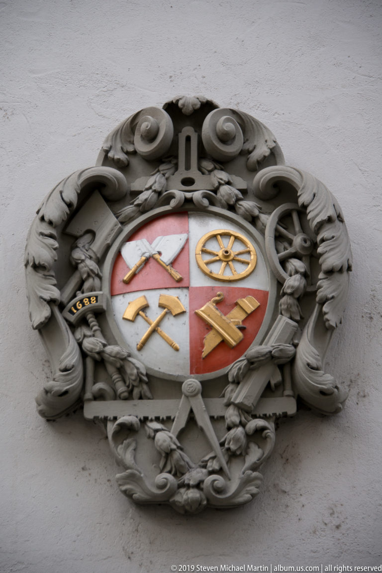 Quartered red-and-white coat of arms with crossed tools and a wheel, framed by carved gray scrollwork.