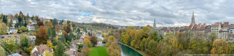 Aare River curving through autumn trees toward Bern rooftops and a tall church spire.