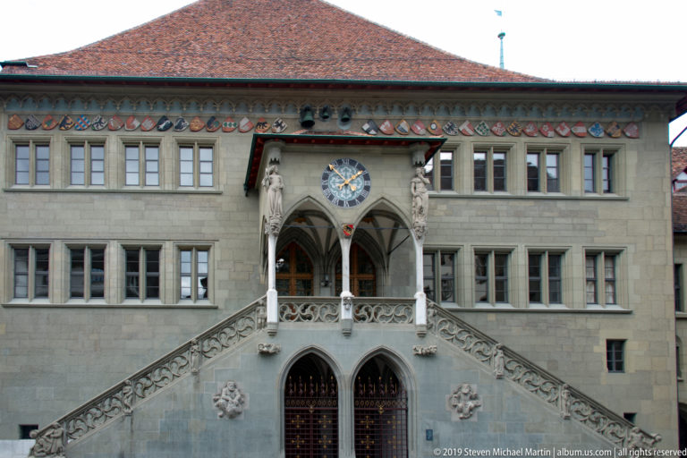 Stone facade of Bern Rathaus with double staircase, pointed arches, clock, and heraldic shields beneath a red tile roof.