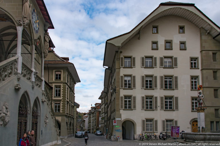 Historic gabled building with shuttered windows at a Bern street corner, beside part of the Rathaus and a distant fountain figure.